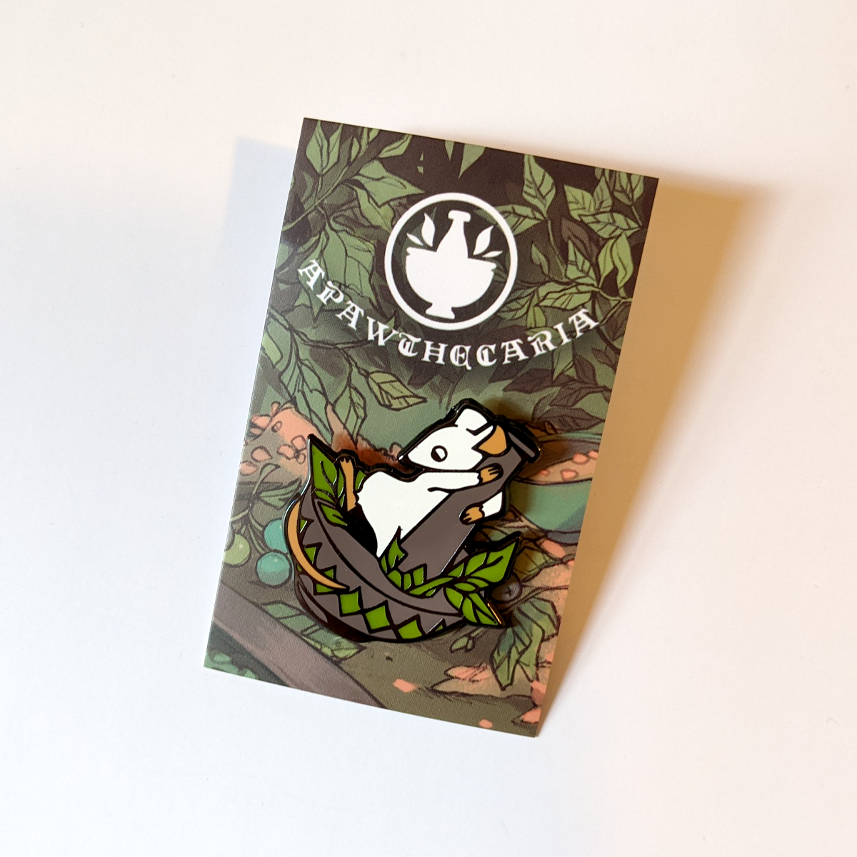 Photo. A smooth enamel pin badge, stuck to a backing card. The pin is of of a stylised cartoon mouse standing in a mortar and pestle, grinding leaves. The backing card shows leaves, vegetation, berries and bowls of reagents.