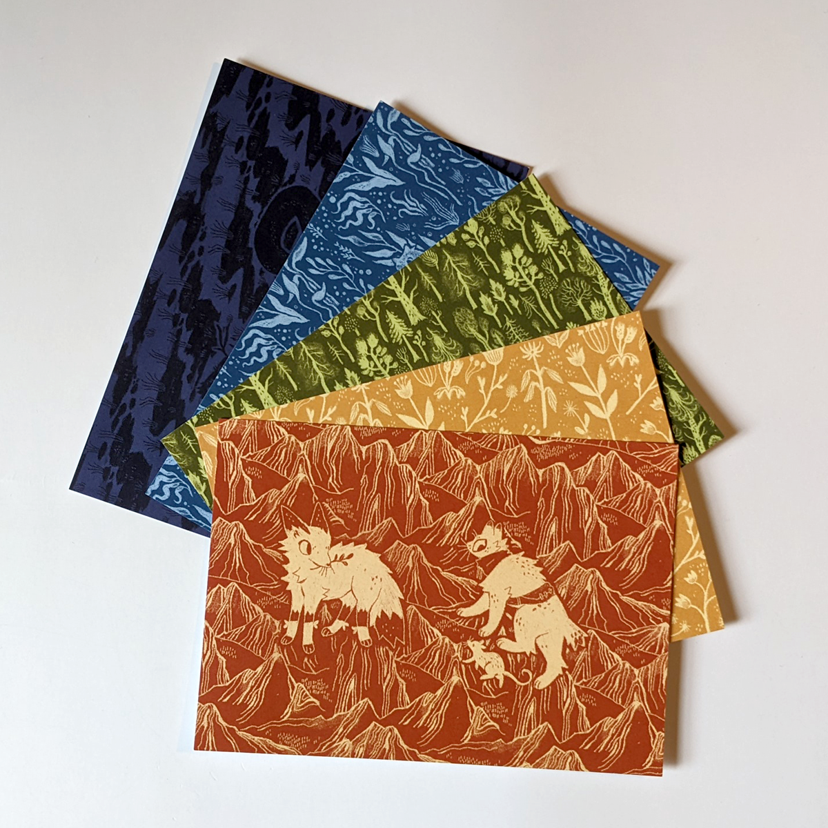 Photo. 5 illustrated postcards featuring patterns from the book Apawthecaria. The cards are themed; red for mountain, featuring a fox glancing at a stout and mouse; yellow for meadows; green for forests; blue for lakes; and purple for bogs.