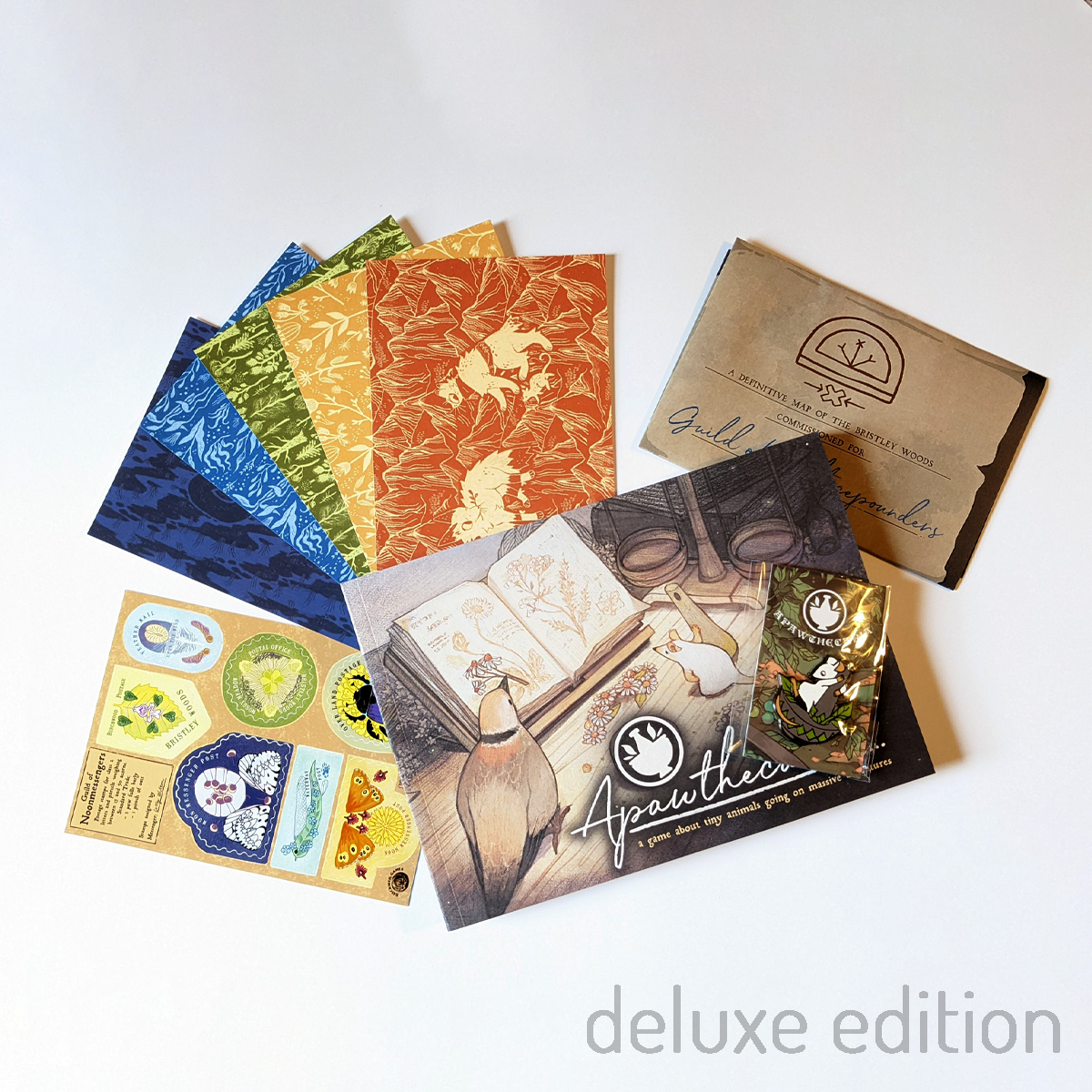 Photo. The deluxe set for Apawthecaria, showing 5 illustrated post cards, a sticker sheet of stamp designs, and a pin badge of a potion-making mouse, alongside the book Apawthecaria and its game map.